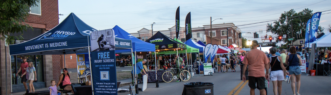 Crowds walk among white vendor tents at an outdoor street fair lined with trees and brick buildings.